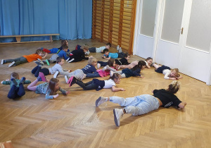 Dzieci leżą na podłodze i wykonują ćwiczenia pokazywane przez prowadzącą warsztaty taneczne.