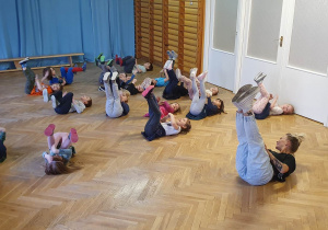 Dzieci leżą na podłodze z nogami uniesionymi do góry i wykonują ćwiczenia pokazywane przez prowadzącą zajęcia taneczne.