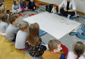Dzieci siedzą w półkolu na dywanie i słuchają prowadzącej warsztaty LABOLO, która wyjaśnia im, o co chodzi z ciśnieniem.