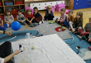 Dzieci siedzą w kole na dywanie i obserwują prowadzącą wykonującą doświadczenie związane z ciśnieniem.