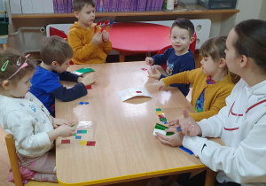 Dzieci siedzą przy stolikach i z pomocą prowadzących przyklejają kolorowe kawałki folii na przezroczystej folii tworząc z nich witraż.