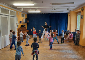 Dzieci stoją w rozsypce na sali gimnastycznej i powtarzają ruchy taneczne za trenerką zumby rozkładając rączki do boku.