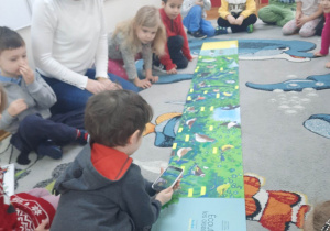 Akcja "Czytamy, głowy otwieramy", dzieci siedzą na dywanie oglądając ilustracje w książeczce, a jeden chłopiec skanuje telefonem ilustracje ptaszka.