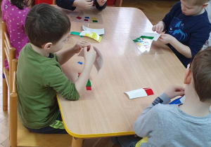 Dzieci siedzą przy stolikach, na przezroczystej folii przyklejają kolorowe kawałki folii tworząc z nich witraż.