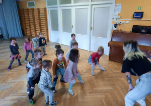 Dzieci stoją w rozsypce na sali gimnastycznej i powtarzają ruchy taneczne za trenerką zumby stojąc w rozkroku z ugiętymi kolanami..