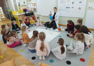 Dzieci siedzą w półkolu na dywanie i obserwują prowadzącą wykonującą doświadczenie związane z ciśnieniem.