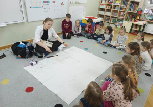Dzieci siedzą w półkolu na dywanie i obserwują prowadzącą wykonującą doświadczenie związane z ciśnieniem.