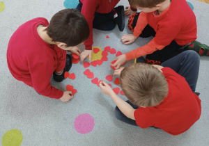 Chłopcy ubrani na czerwono układają duże serce z małych papierowych serduszek.