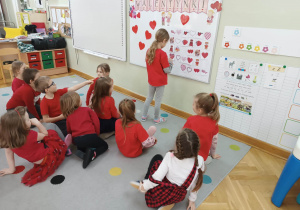 Dzieci siedzą na dywanie, a dziewczynka przy tablicy stara się ułożyć sudoku z obrazków z symbolami miłości.