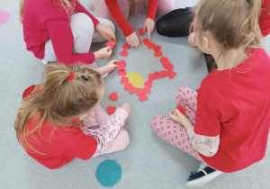 Dziewczynki ubrane na czerwono układają duże serce z małych papierowych serduszek.