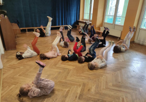 Dzieci stoją w rozsypce na sali gimnastycznej i powtarzają ruchy taneczne za trenerką zumby leżąc na podłodze i podnosząc nogi do góry..