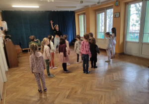 Dzieci stoją w rozsypce na sali gimnastycznej i powtarzają ruchy taneczne za trenerką zumby.