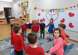 Dzieci ubrane na czerwono tańczą w kole, a w środku stoi dziewczynka z maskotką wielkiego czerwonego serca.