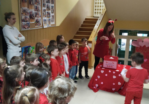 Dzieci ubrane na czerwono stoją i czekają na otrzymanie swojej walentynki, a nauczycielka wyjmuje kolejne.