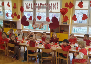 Dzieci ubrane na czerwono siedzą przy stolikach na tle napisu Walentynki i zajadają się pysznościami przygotowanymi z okazji Walentynek.