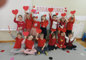 Dzieci ubrane na czerwono z papierowymi serduszkami w rękach pozują do grupowego zdjęcia na tle napisu Walentynki.