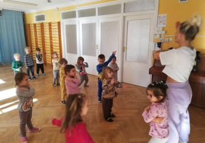 Dzieci stoją w rozsypce na sali gimnastycznej i powtarzają ruchy taneczne za trenerką zumby.