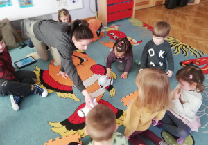 Dzieci siedzą na dywanie, a prowadząca warsztaty z robotyki pokazuje im, jak należy się przywitać z robocikiem.