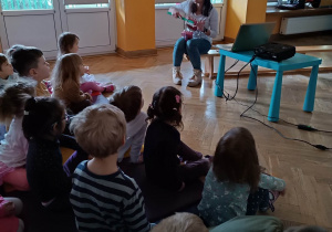 Dzieci siedzą na materacach, a prowadząca warsztaty stomatologiczne pokazuje im jak prawidłowo szczotkować zęby szczoteczką na dużym modelu jamy ustnej.