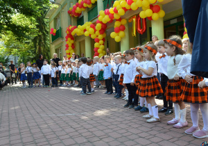 Dzieci z młodszych grup stoją w rzędzie i śpiewają.